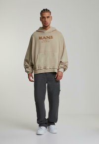 Haut de sweatshirt beige surdimensionné avec une poche avant et un logo imprimé, associé à un pantalon gris foncé et des baskets blanches, se tenant contre un fond neutre.