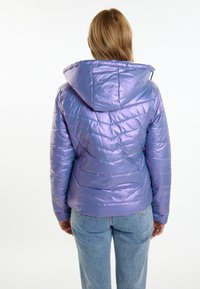 Lilla puffy jakke med hætte, der har en skinnende, quiltet tekstur og skrå syninger. Båret med lyseblå jeans.