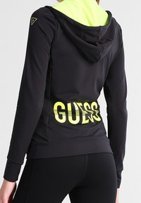 Haut de sport noir avec capuche jaune fluo et graphique au dos. Comprend un logo, des manches longues et un design ajusté. Matière douce et extensible.