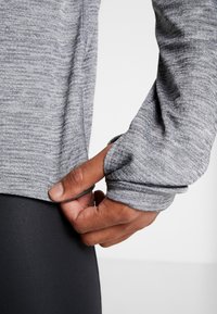 Gray long-sleeve athletic top with textured fabric, fitted cuffs, and side slit. Hand gripping lower hem. Black leggings visible underneath.