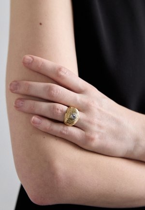 Gold ribbed ring featuring a central clear stone, worn on a fingers crossed over an arm with a smooth texture, and a neutral skin tone.