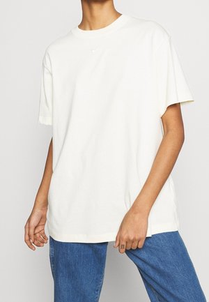 T-shirt en coton écru à coupe décontractée, manches courtes, avec un petit logo Nike au centre de la poitrine, associé à un jean bleu.