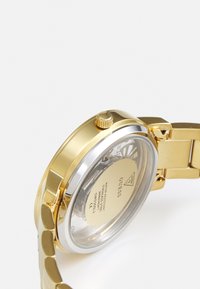 Gold stainless steel watch with a partially transparent back, featuring a silver bezel and a textured gold strap. Includes a crown on the side.