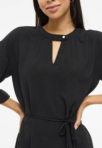 Czarna sukienka z fakturowanego materiału, z dekoltem typu keyhole, krótkimi rękawami i wiązaniem w talii. Minimalistyczny design z delikatnym akcentem w postaci guzika.