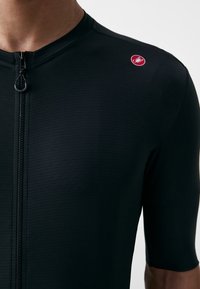 Maglia da ciclismo nera con maniche corte, tessuto a trama, zip frontale e un piccolo logo rosso sulla spalla sinistra.