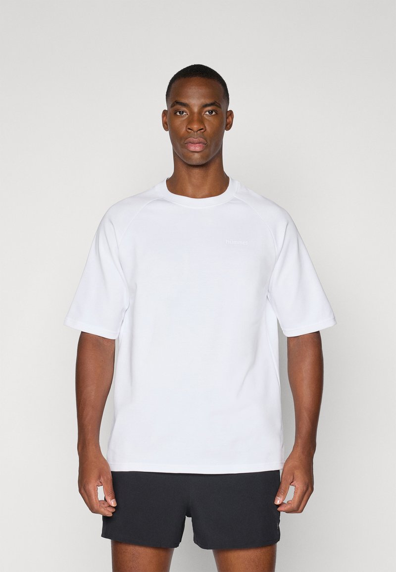Camiseta blanca de manga corta con cuello redondo, hecha de una tela suave. Presenta un logo sutil en gris claro en la parte delantera. Combinada con pantalones cortos negros.