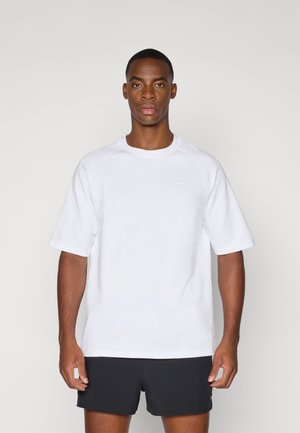 Biała, krótka koszulka z krótkim rękawem o okrągłym dekolcie, wykonana z gładkiego materiału. Z przodu znajduje się subtelne logo w jasnoszarym kolorze. Połączona z czarnymi szortami.