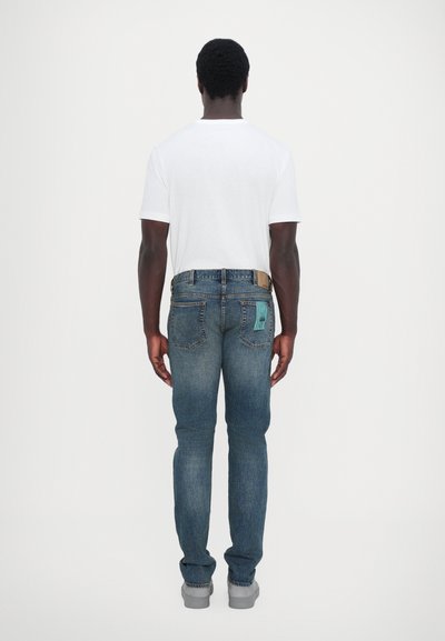 Jean en denim bleu à jambe droite avec une poche arrière ornée d'une touche turquoise, associé à un t-shirt blanc uni et des chaussures grises.