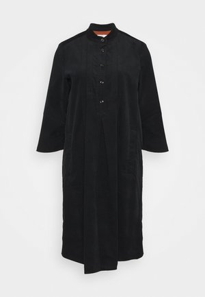 Mustan pitkä takki, jossa on mandariinikaulus, edessä nappikiinnitys, kaksi sivutaskua ja sileä, tekstuurihohtoinen kangas.