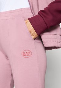 Hånd i lommen på lyserosa bukser med EA7 Emporio Armani-logo, iført en burgunderrød genser med lange ermer, nærbilde.