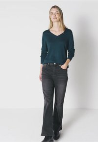 Maglione verde scuro a scollo a V con maniche lunghe, abbinato a jeans neri a zampa d'elefante. La modella sta in piedi con una mano nella tasca, indossando stivali neri alla caviglia.