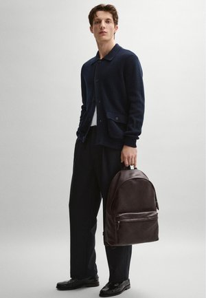 Giovane uomo in piedi, indossa un cardigan blu scuro, pantaloni neri a gamba larga, mocassini neri, tiene uno zaino in pelle marrone scuro per la maniglia.