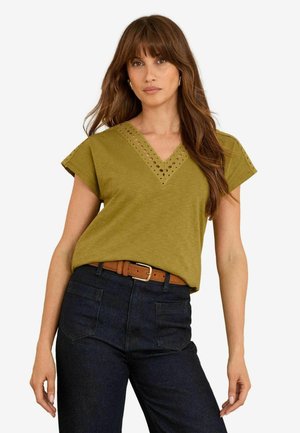 Vrouw met lang bruin haar, gekleed in een olijfgroene V-hals top met oogjesdetails en donkerblauwe jeans met een bruine riem, staand tegen een witte achtergrond.