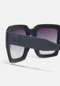 Črne plastične sončna očala z velikanskimi angularnimi okvirji, vijoličnimi gradientnimi lečami in izrazitim logotipom "CK" na strani.