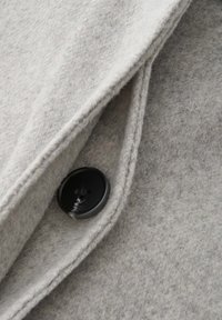 Close-up of light gray wool fabric with folded edges and a black four-hole button sewn onto the material.