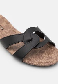Sandale marron à motif avec des lanières en cuir noir qui se croisent sur le dessus et un détail découpé circulaire près de la zone des orteils.