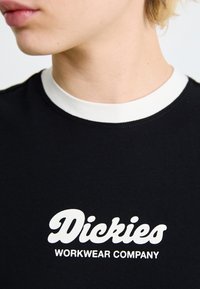 Čierne tričko s bielym okrúhlym výstrihom, s výrazným bielym textom "Dickies WORKWEAR COMPANY" na prednej strane. Tkanina sa zdá byť hladká.