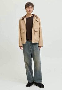 Young person standing wearing a beige jacket, black shirt, loose blue jeans, and black shoes against a plain light background.