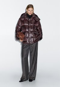 Brown puffer jacket with a shiny finish, large pockets, a high collar, and a cinched waist, paired with gray trousers and a brown clutch.