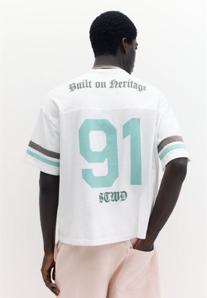 Homme à la peau foncée portant une chemise blanche avec le numéro turquoise 91, le texte « Built on Heritage » et des manches rayées, debout avec la main dans la poche d’un pantalon rose.