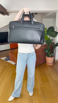 Fekete textúrájú laptop táska dupla fogantyúval és cipzárral záródik. Diszkrét logóval és elegáns dizájnnal rendelkezik, sima felülettel kiegészítve.