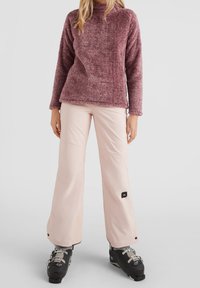 Puszysty bordo sweter z wysokim kołnierzem, zestawiony z jasnoróżowymi, szerokimi spodniami narciarskimi. Czarne buty narciarskie z metalowymi klamrami.