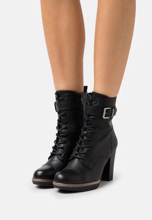 Stivali con tacco alto in pelle nera, alla caviglia, con lacci, zip laterale e cinturino, indossati su gambe nude su uno sfondo bianco uniforme.