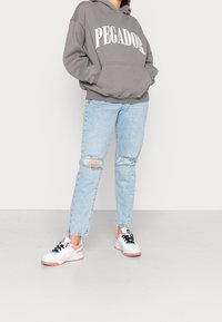 Grauer Kapuzenpullover mit weißem Schriftzug "PEGADOR", kombiniert mit hellblauen zerrissenen Jeans und weißen Turnschuhen mit rosa Akzenten.