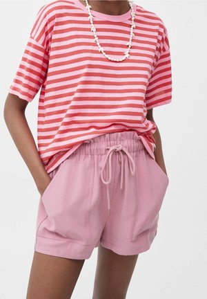 Person trägt rosa und rot gestreiftes kurzärmeliges Hemd, rosa Shorts mit Kordelzug und eine weiße Perlenkette, Hände in den Taschen.
