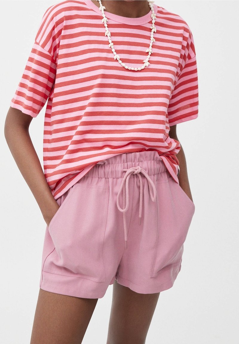 Personne portant une chemise à manches courtes rayée rose et rouge, un short rose avec cordon de serrage, et un collier de perles blanches, les mains dans les poches.