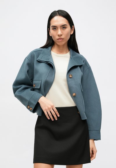 Przycięta niebieska kurtka z zapięciem na guziki, dużym kołnierzem i dwiema kieszeniami na piersi. Noszona na beżowej bluzce i czarnej spódnicy.