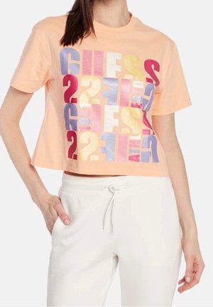 Nainen, jolla on persikanvärinen lyhyt t-paita, jossa monivärinen GUESS-logo, ja valkoiset naruviikselliset housut, käsi taskussa, yksiväristä taustaa vasten.