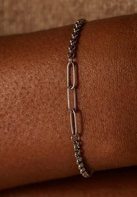 Zilveren armband met ovale schakels en een gestructureerde afwerking, liggend op een bruine huidskleur, met duidelijke gepolijste en matte secties.
