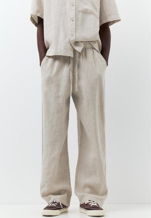 Muž oblečený v béžových ľanových nohaviciach so sťahovacou šnúrou a zodpovedajúcej krátkorukávovej košeli s gombíkmi, s rukami vreckách, doplnený tmavými teniskami s bielymi podrážkami.