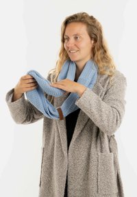 Lichtblauwe gebreide infinity sjaal vastgehouden door een persoon, met een gestructureerd patroon en een bruine leren label. De jas is grijs en fuzzy.