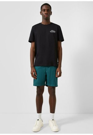 Junger Mann steht frontal, trägt ein schwarzes T-Shirt mit weißem Text, türkisfarbene Shorts, weiße Socken und cremefarbene Sneakers vor einfachem Hintergrund.