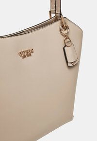 Bolsa tote beige hecha de material sintético suave. Presenta un logo en tono dorado, una etiqueta de bolso y acentos de hardware redondo en la correa.