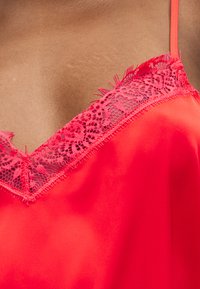 Κοντινή λήψη κόκκινου σατέν ρούχου με διακοσμητικό δαντελένιο λουλουδάτο χείλος που φοριέται στο επάνω μέρος του στήθους και του ώμου ενός ατόμου με ορατό λεπτό ιμάντα.