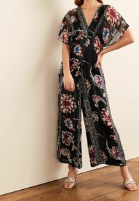 Combinaison noire à fleurs avec jambes larges, arborant des motifs rouges et bleus et un décolleté en V profond. Tissu transparent avec une finition texturée.