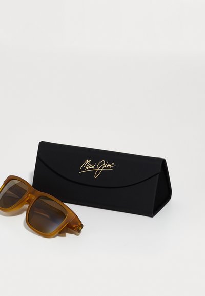 Maui Jim solbriller med overdimensionerede brune linser, gennemsigtig orange ramme og sort beskyttelsescase med guldlogo.
