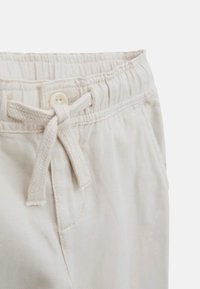 Vaaleanbeige puuvillahousut, joissa on elastinen vyötärö, kiristysnauha, napin suljenta ja sivutaskut. Rakenne pinnassa.