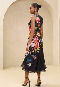 Blumiges Midi-Kleid mit schwarzem Hintergrund, großen bunten Blumen, ärmellosem Design und fließendem Saum. Akzentuiert mit Riemchen-Absätzen.