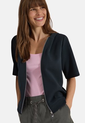Femme souriante, portant une veste noire zippée à manches courtes par-dessus un haut rose et un pantalon gris avec cordon, mains dans les poches.
