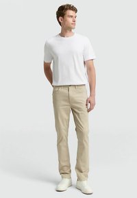 Béžové bavlněné chino kalhoty se střihem rovného střihu, v kombinaci s bílým tričkem s krátkým rukávem. Bílé tenisky. Model stojí na neutrálním pozadí.