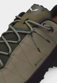 Κοντινό πλάνο πεζοπορικού παπουτσιού σε ελιά και μπεζ με λεπτομερείς κορδόνια, διάτρητη γλώσσα, ετικέτα GORE-TEX και υφή μαύρης σόλας.