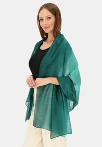 Przezroczysty zielony szal o lekkim, płynnym kroju. Posiada szerokie rękawy i gładką fakturę, elegancko opadający na czarną bluzkę.