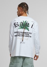 Langärmliges weißes T-Shirt mit schwarzem und grünem Palmenbaum-Graphic, fettgedrucktem Text und einem Etikett mit Details. Weiches Baumwollmaterial, lockere Passform.