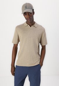 Camisa polo de manga corta en color beige con cuello clásico, combinada con pantalones de lino azules. El conjunto incluye una gorra azul claro con detalles del logo.