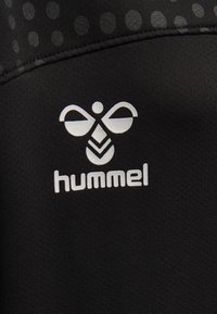 Tessuto nero con una superficie liscia e testurizzata presenta un logo bianco che assomiglia a un'ape, posizionato centralmente, sopra la parola "hummel."