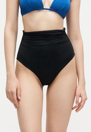 Γυναίκα που φοράει μπλε τοπ μαγιό και μαύρο μαγιό με ψηλή μέση, μπροστά σε απλό ανοιχτό φόντο.